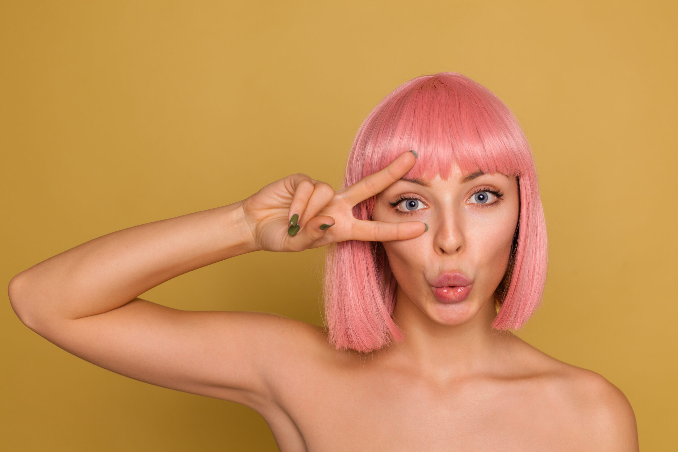 Horizontal shot of pretty young blue-eyed lady with short pink hair holding victory gesture near her face and folding her lips together while posing over mustard background