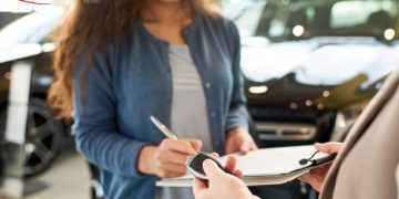Close-up shot of smiling curly woman signing contract for new car, unrecognizable saleswoman holding key in hand, interior of modern showroom on background