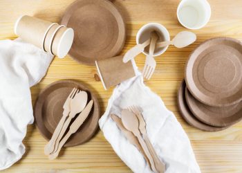 Brown paper cups, plates, wooden cutlery, white linen napkins on wooden background. Recycling concept. Top down view, horizontal image