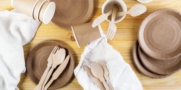 Brown paper cups, plates, wooden cutlery, white linen napkins on wooden background. Recycling concept. Top down view, horizontal image