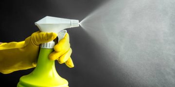 Woman wearing yellow rubber gloves using green spray bottle and spraying liquid mist in air, cool lighting effect. Lot of copy space.